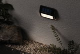 Black solar wall light with warm white LED on gray exterior wall above gravel and plants