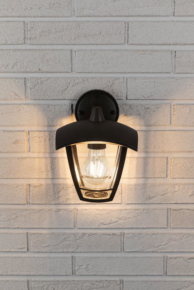 Zwarte metalen buitenwandlamp met heldere glazen kap aan witte bakstenen muur, warme LED-lamp brandt.