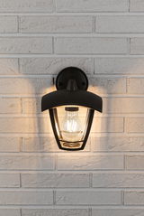 Zwarte metalen buitenwandlamp met heldere glazen kap aan witte bakstenen muur, warme LED-lamp brandt.