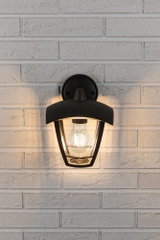 Schwarze Außenwandleuchte aus Metall mit klarer Glashaube an weißer Ziegelwand, LED-Lampe leuchtet warm.