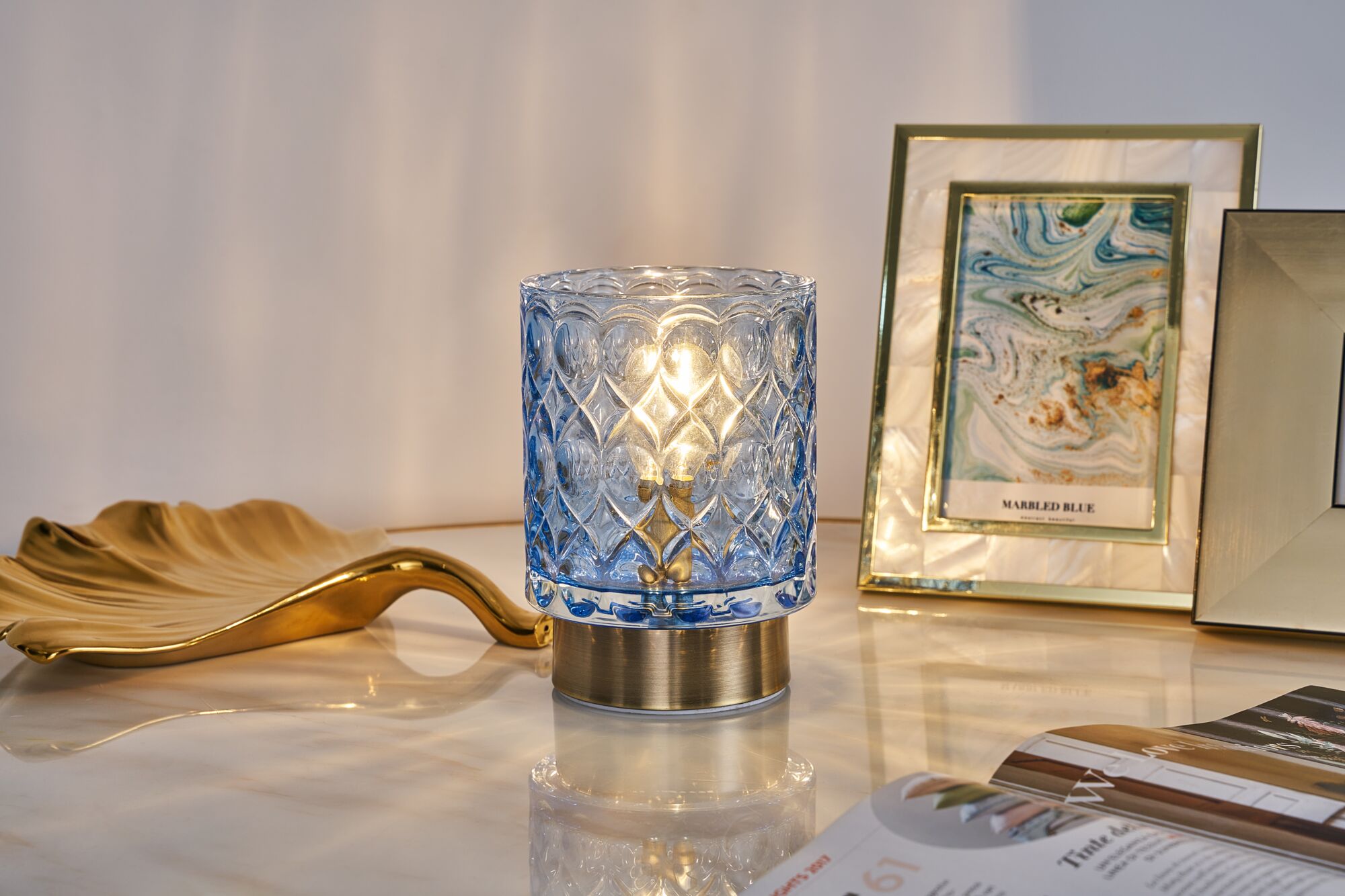 Blue crystal glass table lamp with brass base on marble surface beside gold decor and picture frames