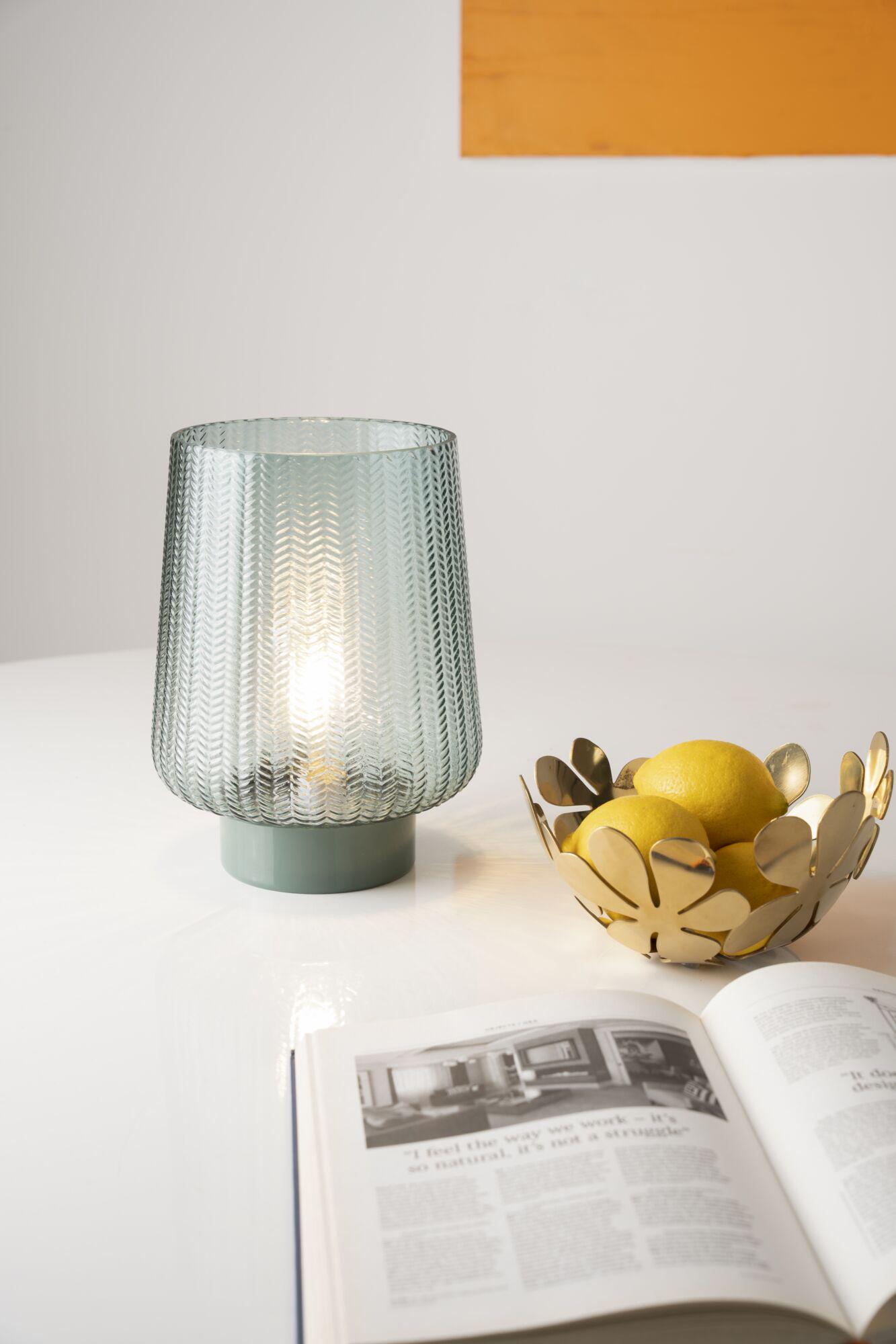 Grøn mønstret glasbordlampe med grå base på hvidt bord ved siden af gylden frugtskål med citroner.