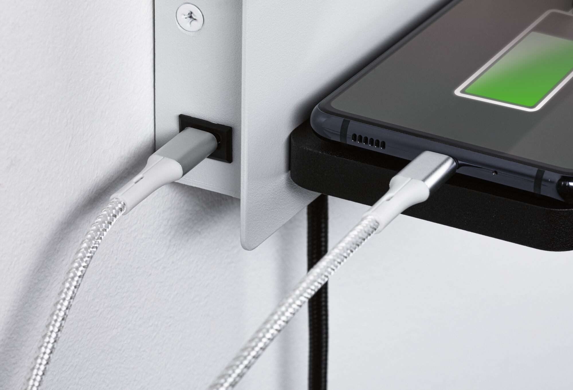 Nahaufnahme eines weißen USB-C Ladekabels mit geflochtener Ummantelung an schwarzem Smartphone und Wandanschluss