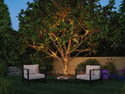 Garten mit Baum, der mit warmweißen LED-Lichterketten umwickelt ist, gemütliche Sitzmöbel und Pflanzen