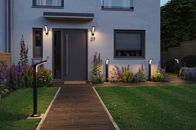 Modern black metal LED outdoor lights illuminate pathway and entrance of house with garden at dusk.