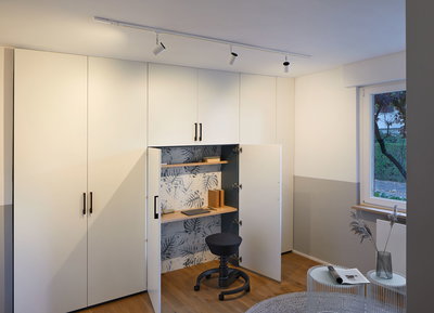 White built-in cabinet with integrated workspace, wooden shelves and black swivel stool in modern room