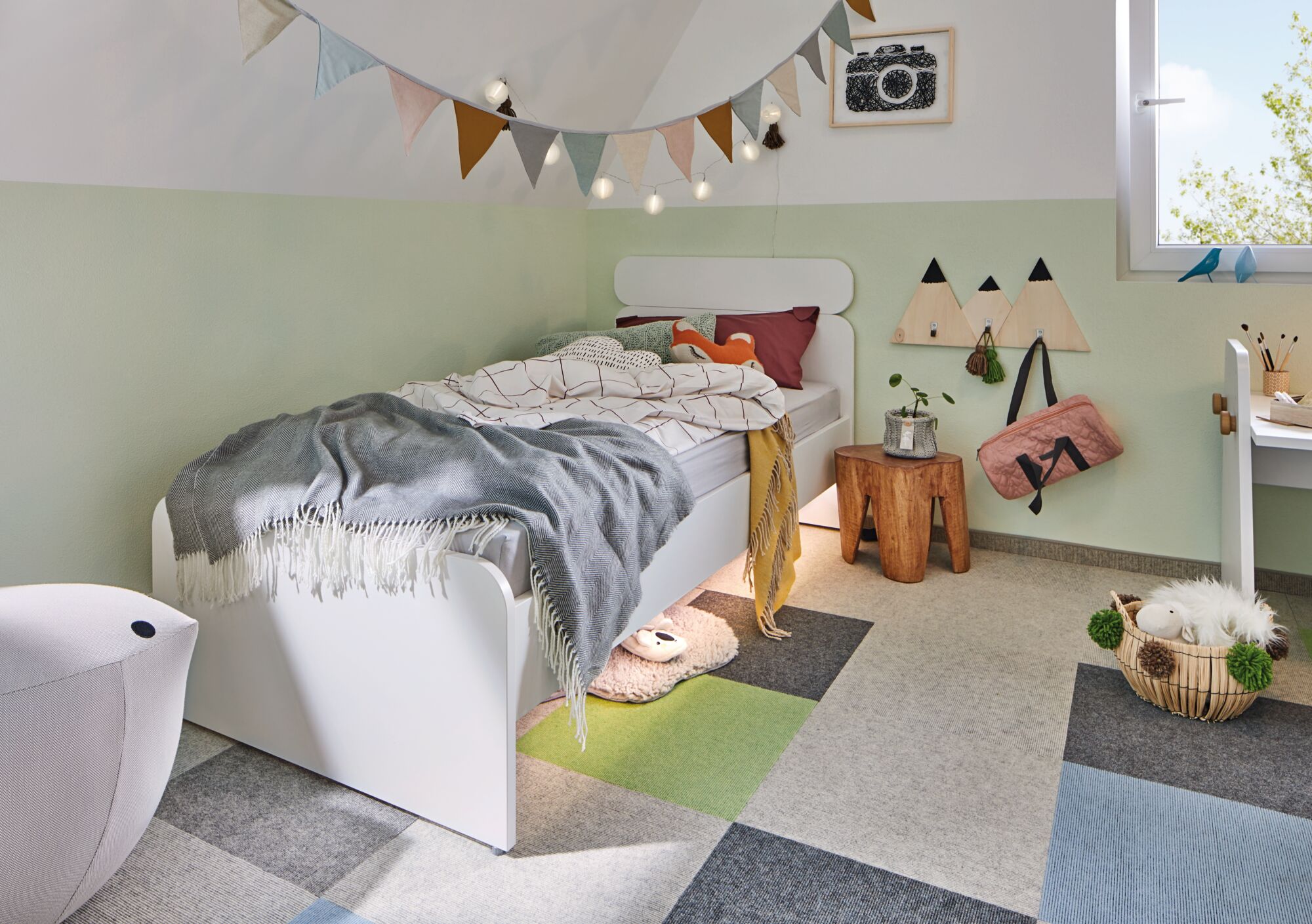 Child's room with white wooden bed, colorful carpet tiles, and decorative fabric bunting on wall