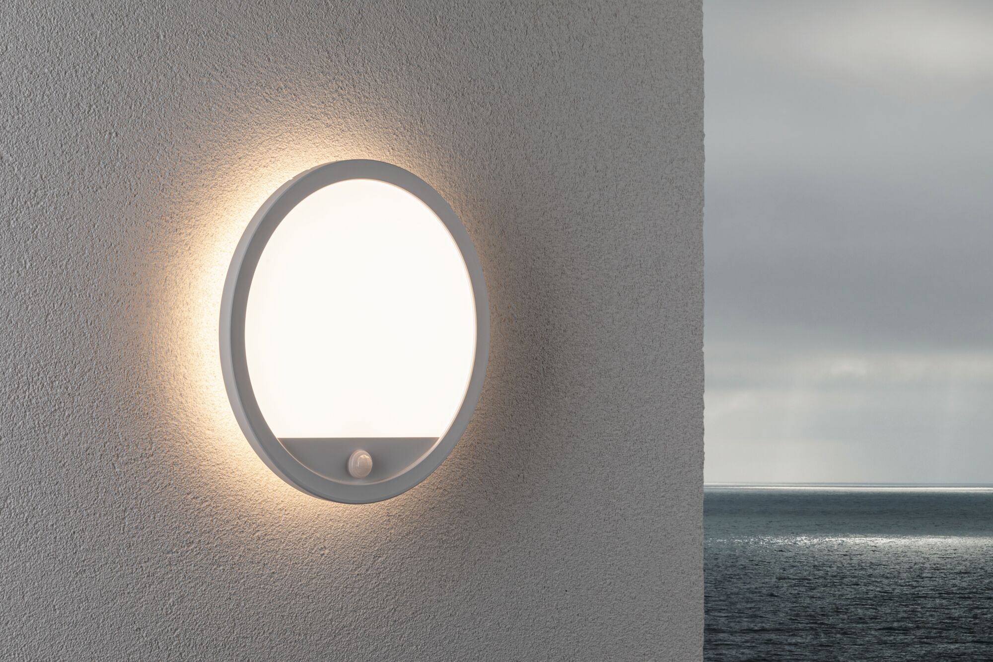 Runde weiße LED-Wandleuchte mit Bewegungsmelder an grauer Wand, Außenbeleuchtung mit Meerblick