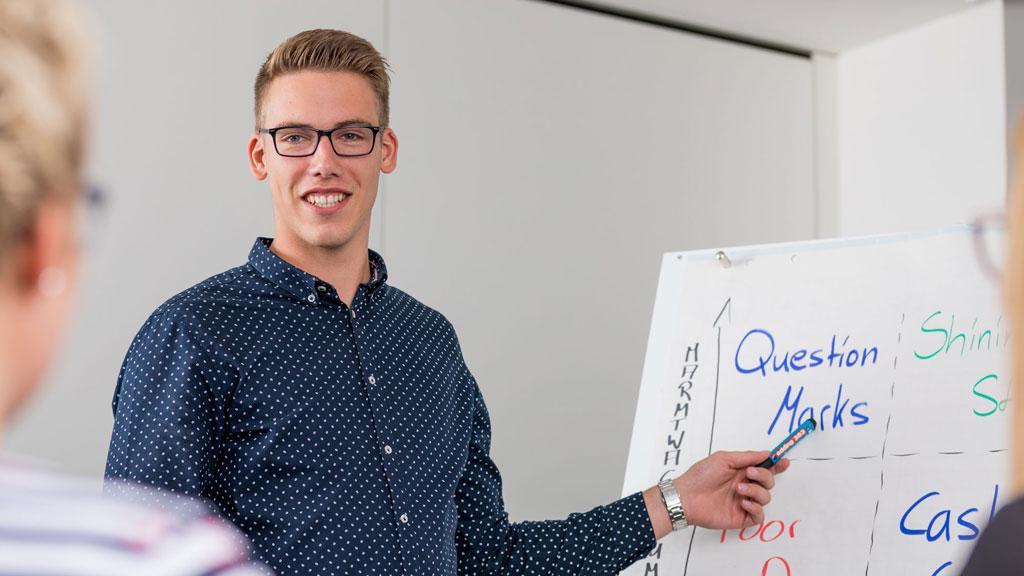 Junger Mann mit Brille erklärt an Whiteboard mit Diagramm und Text in Büro-Meeting.