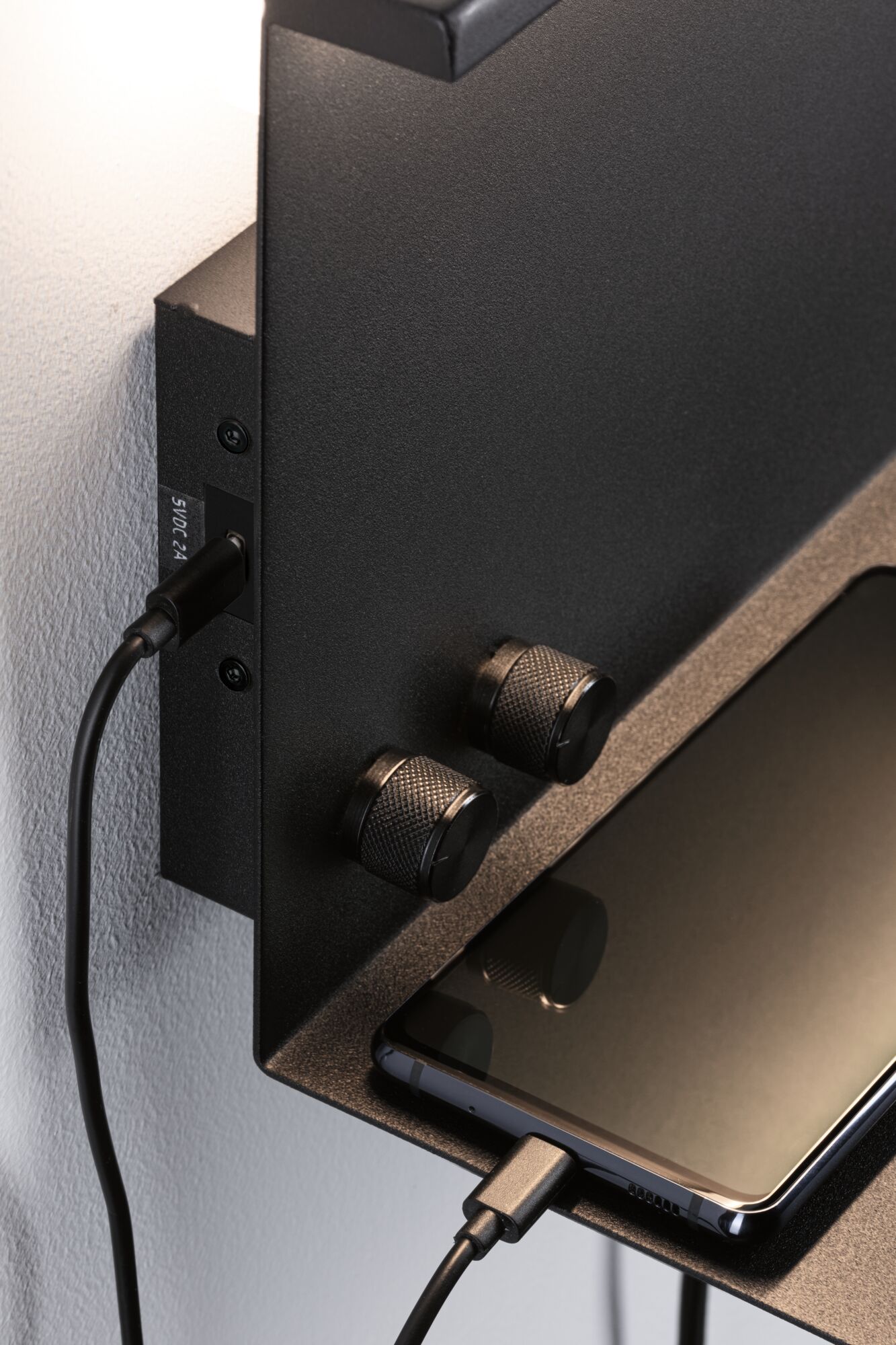 Black metal wall shelf with two textured knobs and smartphone connected via USB charging cable.