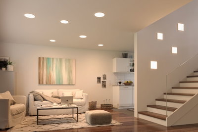 Modern living room with white recessed ceiling lights and square wall lamps by staircase, wooden floor