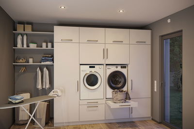 Moderne weiße Einbau-Waschküche mit Waschmaschine, Trockner und integriertem Bügelbrett in hellem Holzraum.