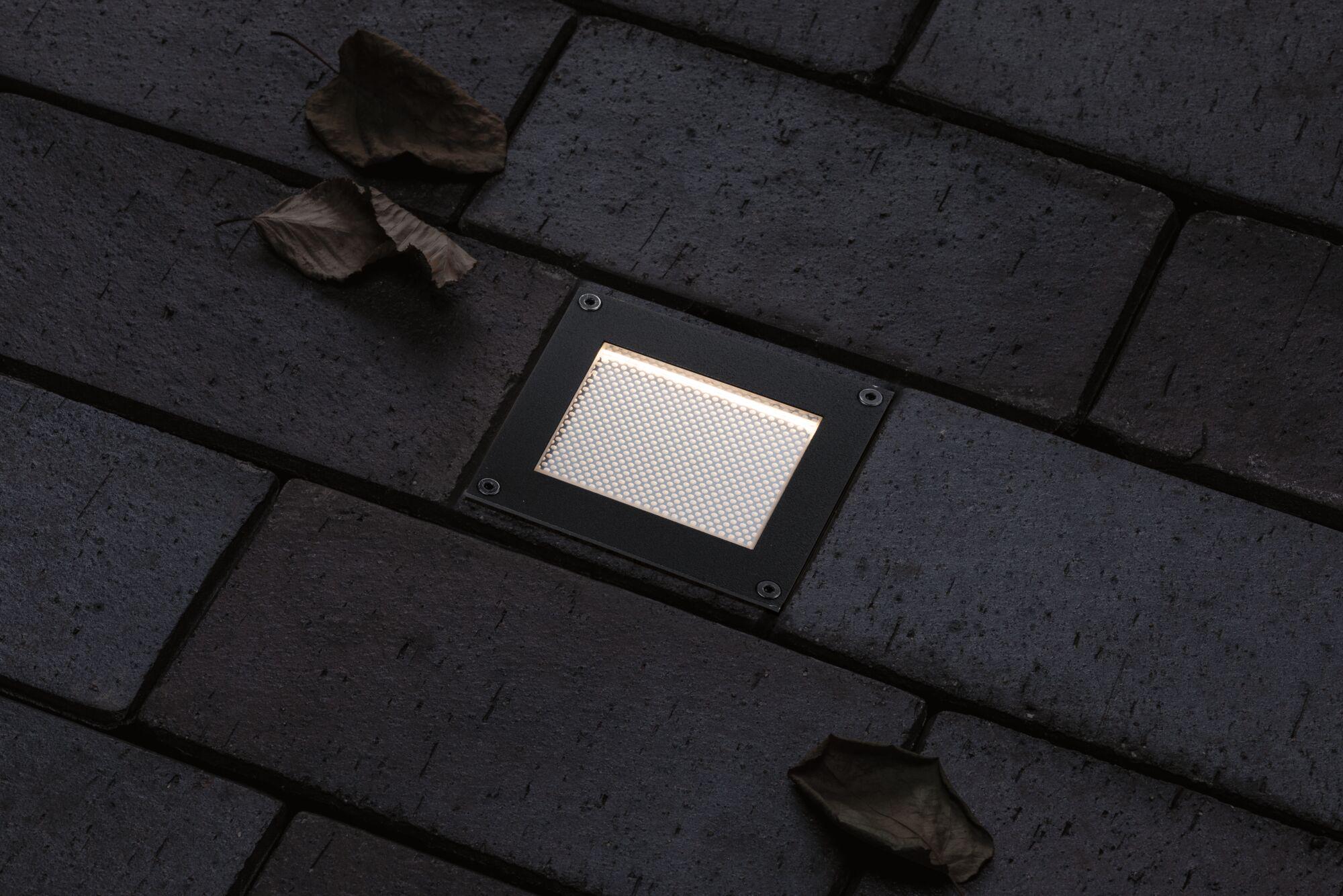 Square black metal LED floor light with warm white illumination on dark pavement surface