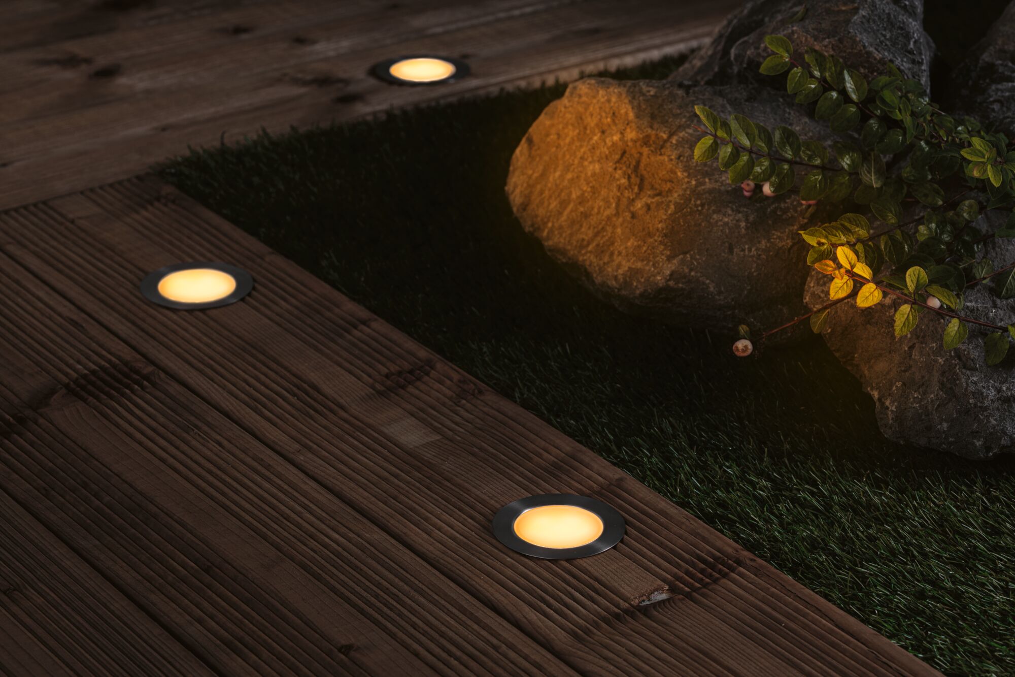 Three recessed LED floor lights with warm white glow on dark wooden deck in garden setting