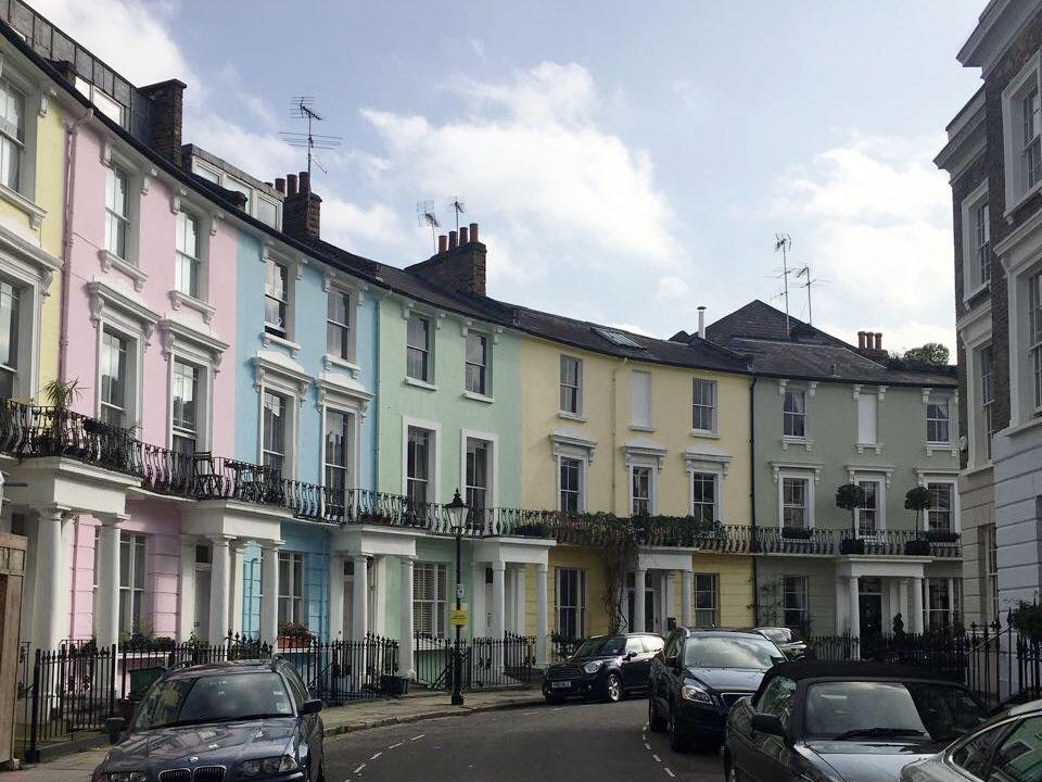 Reihe pastellfarbener Stadthäuser mit weißen Veranden und parkenden Autos an einer ruhigen Straße.