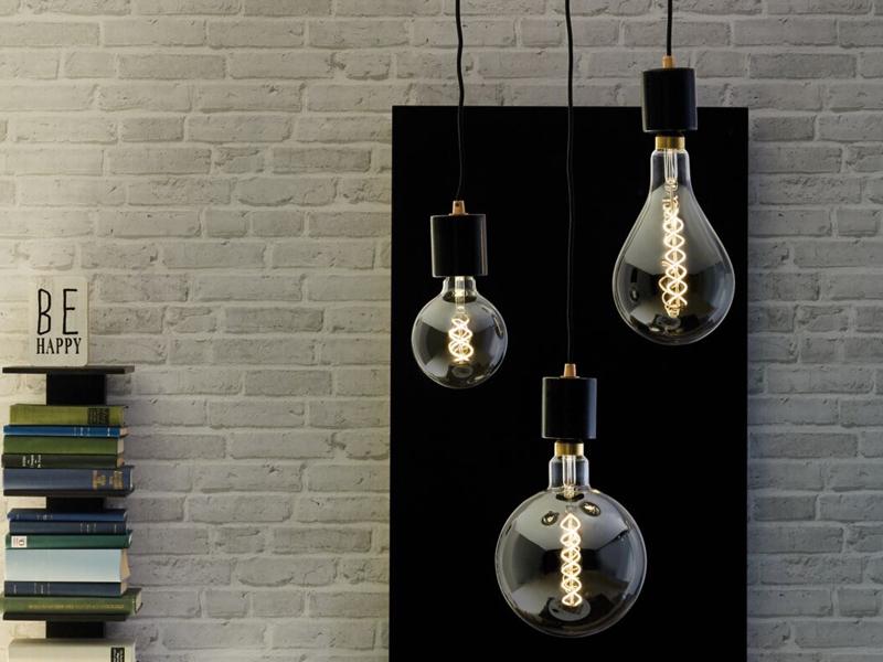 Drei hängende LED-Glühbirnen mit Spiralfaden in klarem Glas vor grauer Ziegelwand und Bücherstapel.