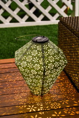 Green solar lantern with floral metal pattern on wooden deck, decorative outdoor garden lighting.