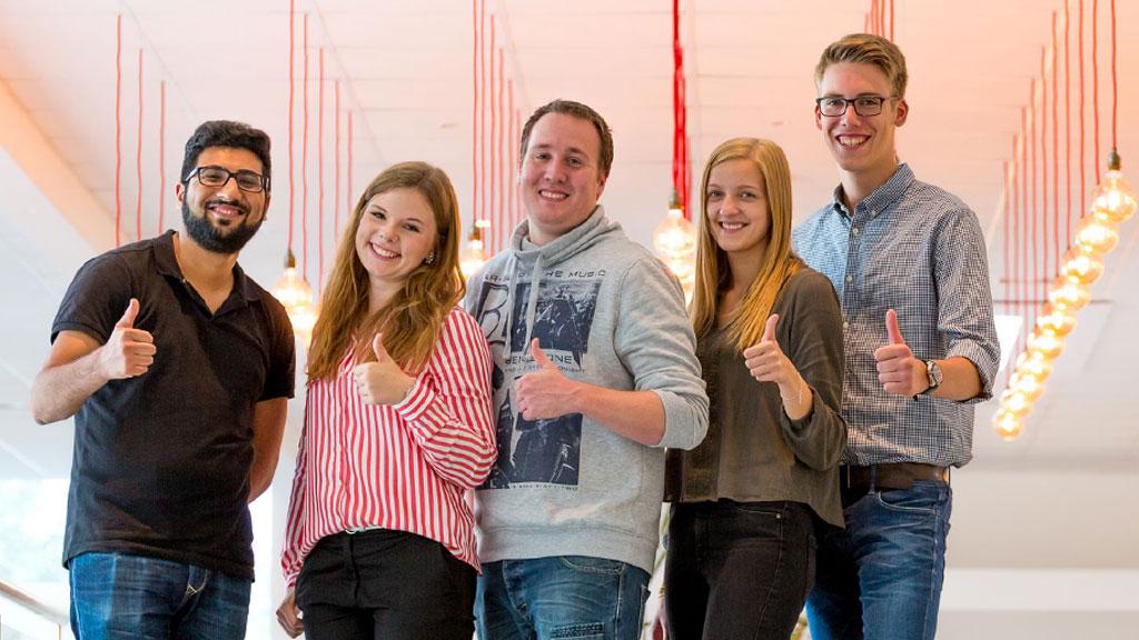 Fünf junge Menschen in moderner Umgebung zeigen Daumen hoch, symbolisieren Teamgeist und Karrierechancen.