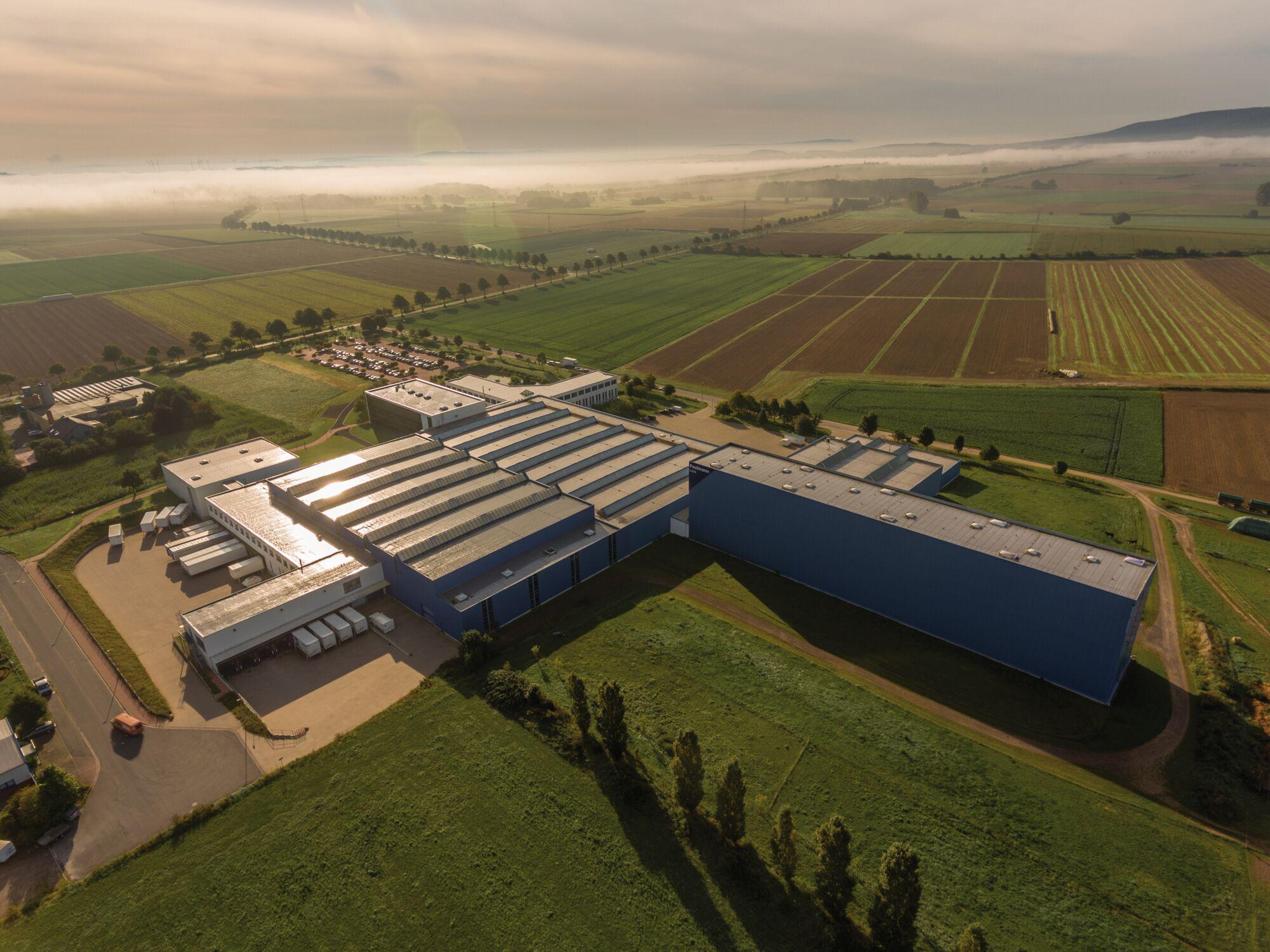 Luftaufnahme eines großen blauen Industriegebäudes umgeben von grünen Feldern und Parkplätzen bei Sonnenaufgang