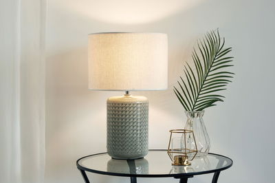 Grey ceramic table lamp with white fabric shade on glass table next to glass vase with green leaf