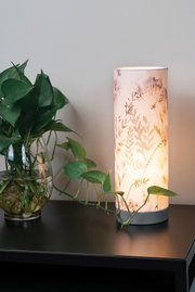Zylindrische Tischlampe mit floralem Stoffschirm in Beige auf schwarzem Tisch neben grünem Pflanzenblatt