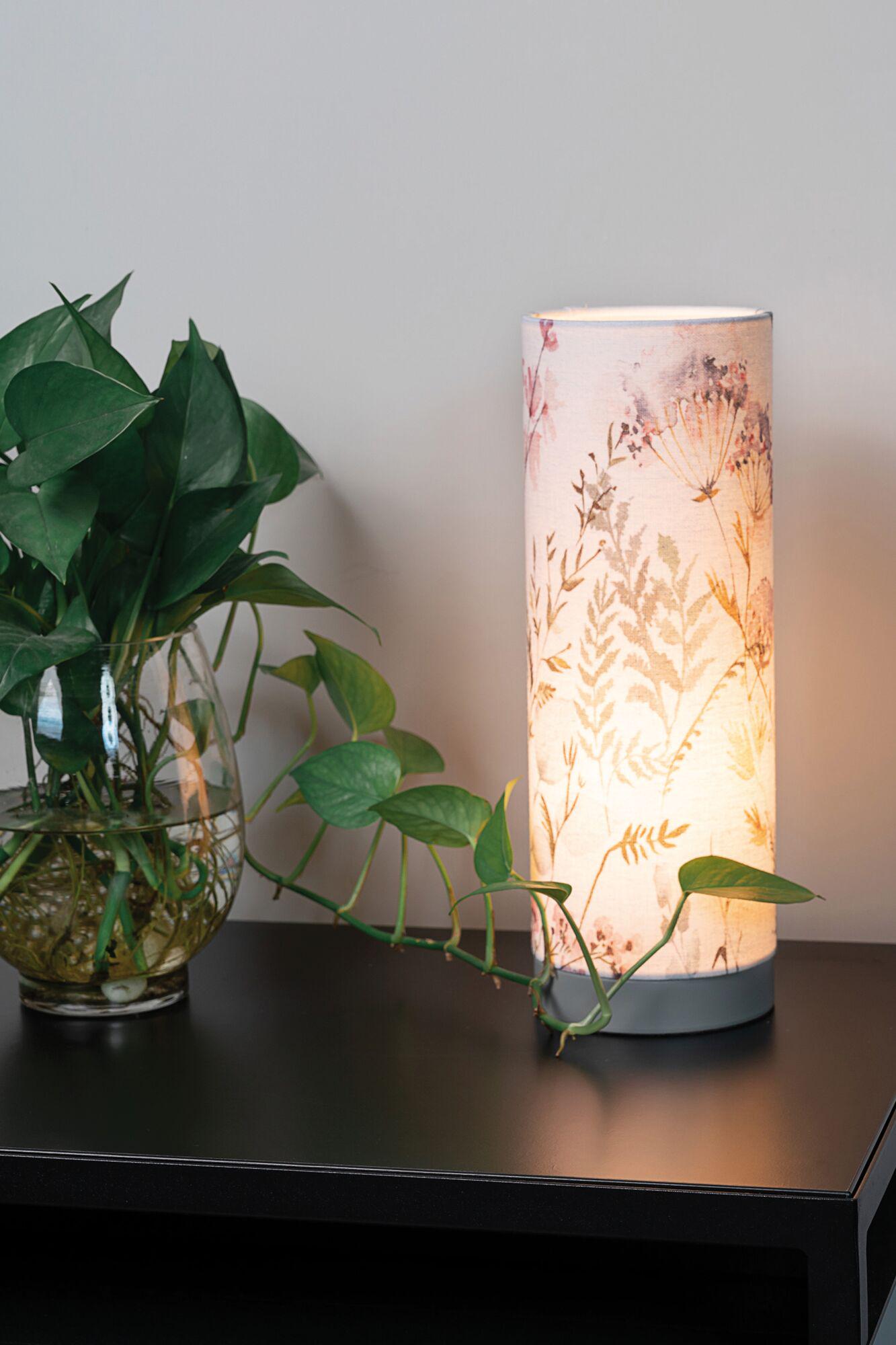 Zylindrische Tischlampe mit floralem Stoffschirm in Beige auf schwarzem Tisch neben grünem Pflanzenblatt