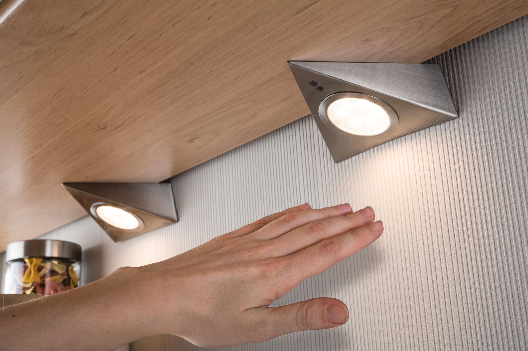 Triangular brushed stainless steel LED under-cabinet lights with warm white light on wood surface.