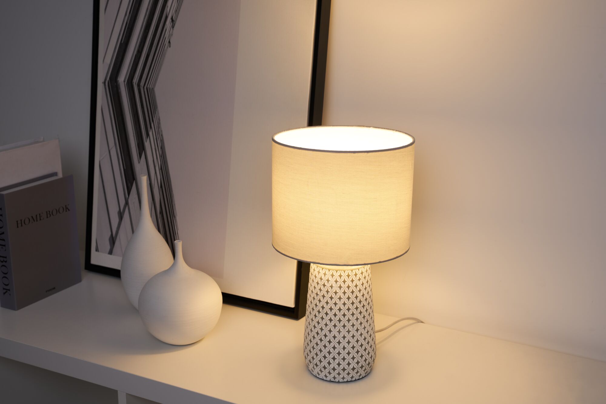 Table lamp with white fabric shade and patterned ceramic base on white shelf with decor items.