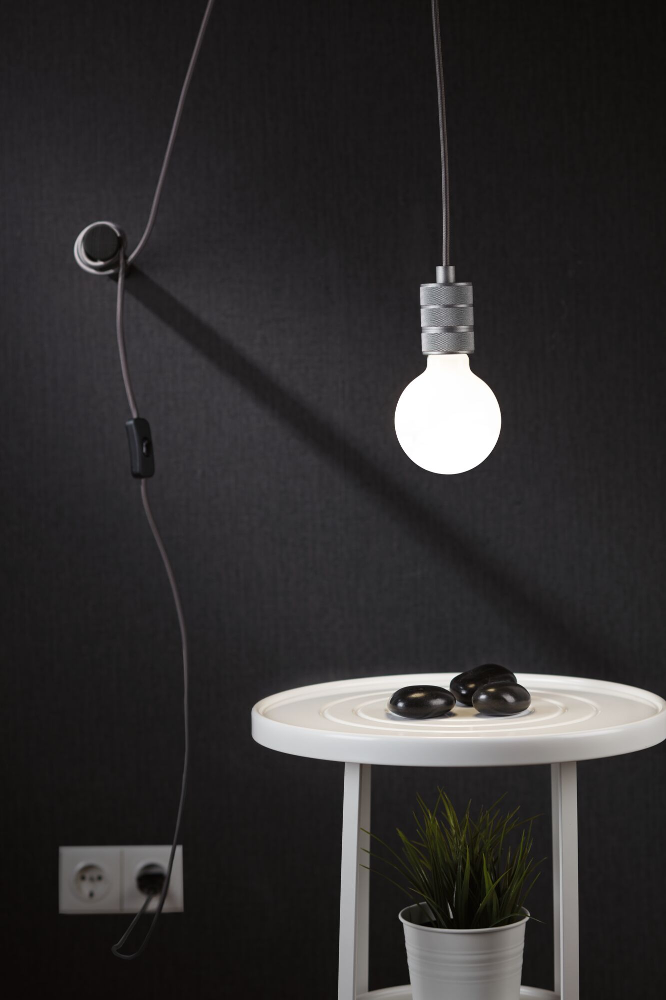 Hængende LED-pære med gråt kabel mod sort væg over hvidt sidebord med plante og sorte sten