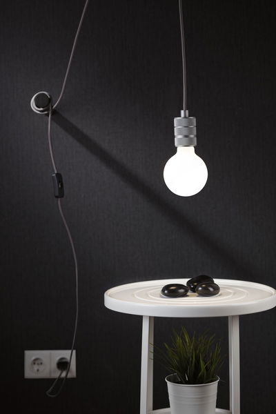 Hængende LED-pære med gråt kabel mod sort væg over hvidt sidebord med plante og sorte sten