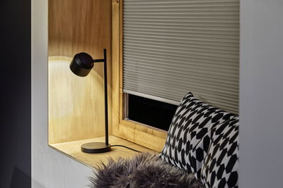 Black metal desk lamp with round head on wooden windowsill near patterned cushion and pleated blind.