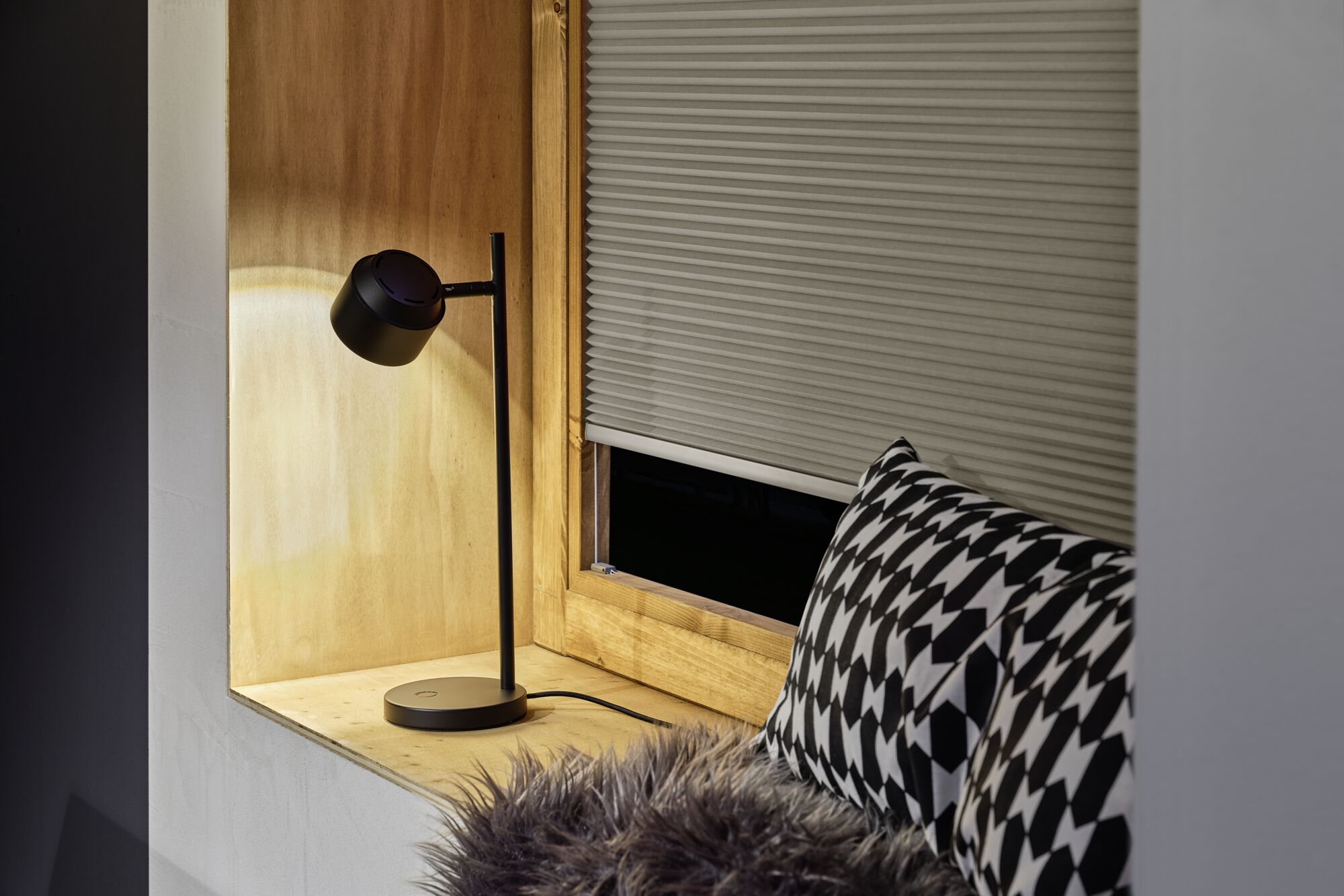 Zwarte metalen bureaulamp met ronde kap op houten vensterbank naast kussen met patroon en plissé gordijn.
