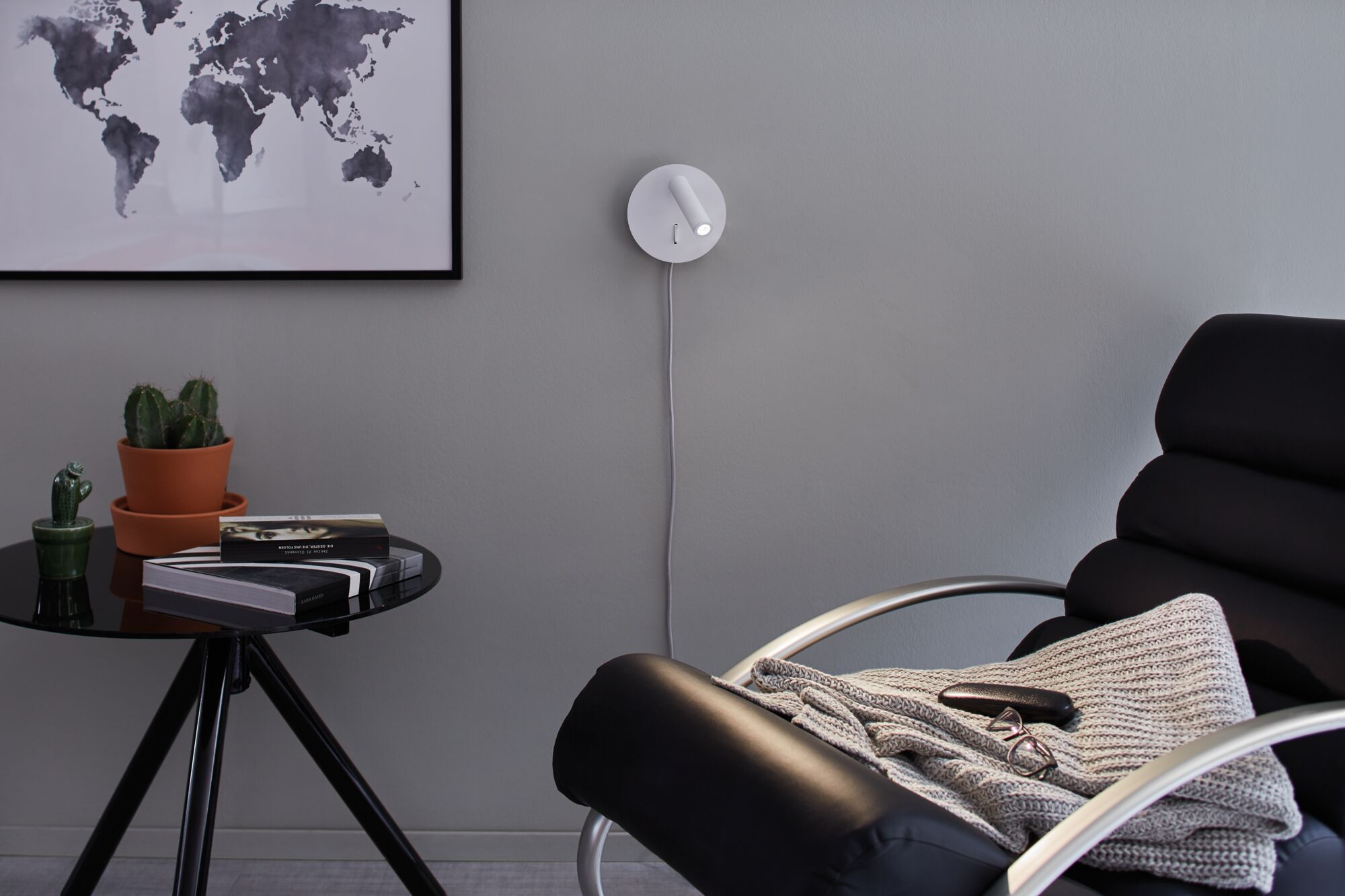 Moderner weißer LED-Wandleuchter mit rundem Sockel an grauer Wand im Wohnzimmer mit schwarzem Sessel.