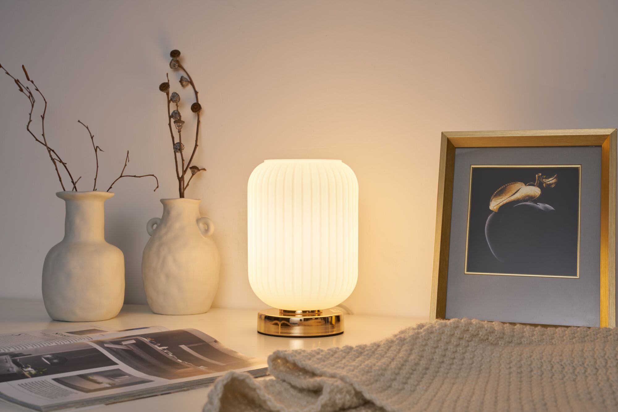 Modern white table lamp with ribbed glass and gold base beside ceramic vases and framed artwork