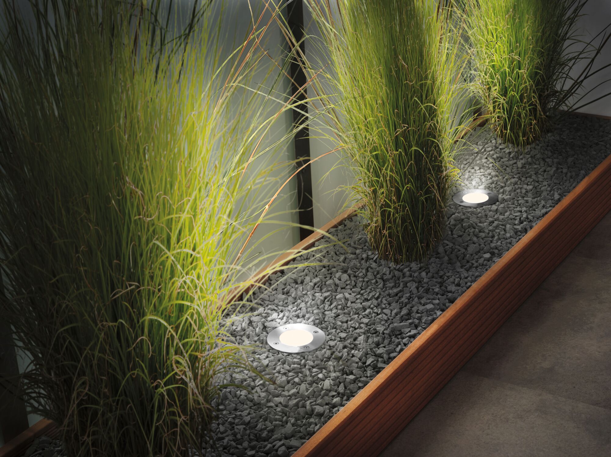 Luminaires LED encastrés en acier inoxydable éclairant des plantes vertes dans un lit de gravier avec cadre en bois