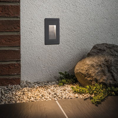 Black metal LED wall light on white wall illuminating pebbles and plants outdoors