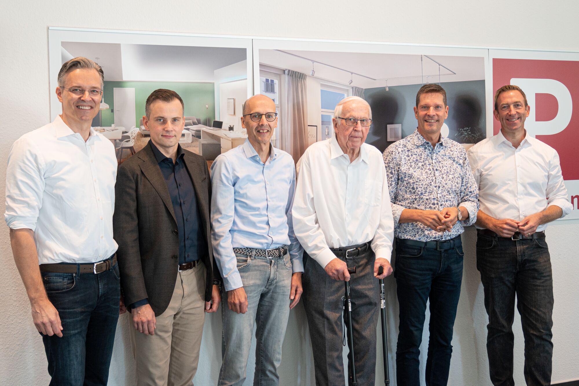 Six men in business attire standing in front of wall with pictures, formal farewell scene in office setting.