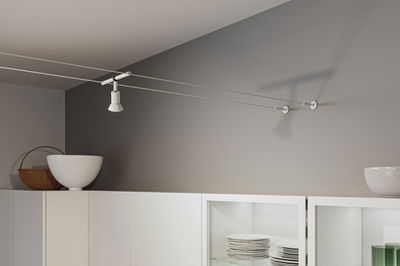 White metal cable spotlight on grey wall above white kitchen cabinet with dishes and decor.