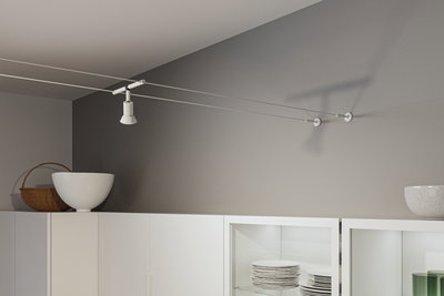 Witte metalen kabelspot aan grijze muur boven witte keukenkast met servies en decoratie.