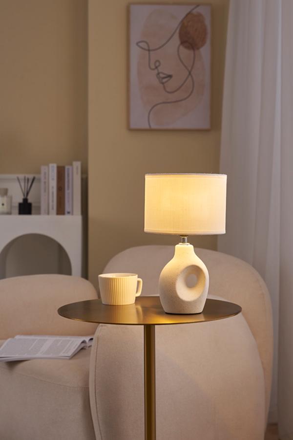 Beige table lamp with round ceramic base and light fabric shade on metal table beside armchair in living room.