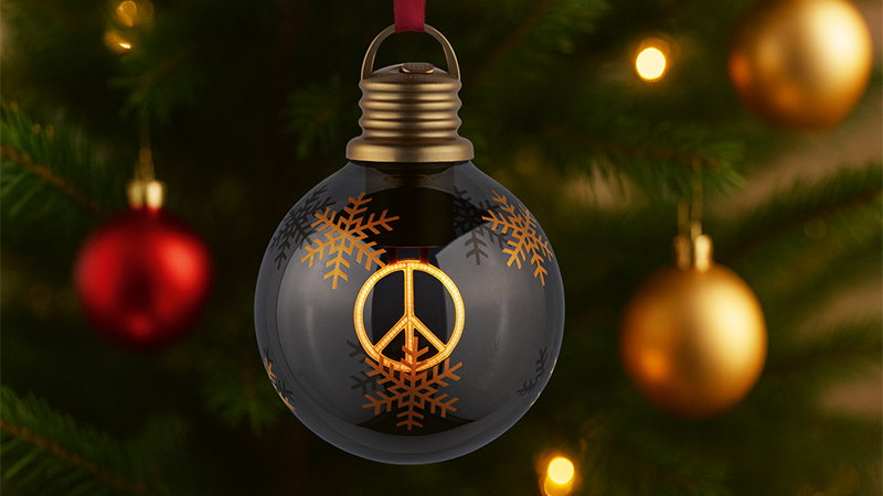 Black glass Christmas bauble with golden peace sign and snowflakes hanging on pine branch.