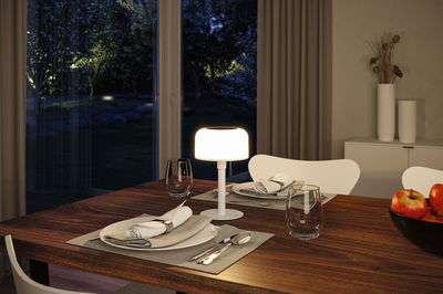 Modern white table lamp with warm white light on wooden table near set dining area and apples