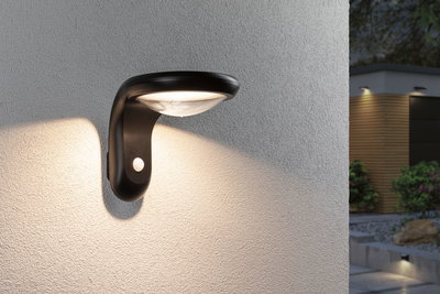 Zwarte buitenwandlamp van kunststof met bewegingssensor verlicht huismuur en tuinpad 's nachts