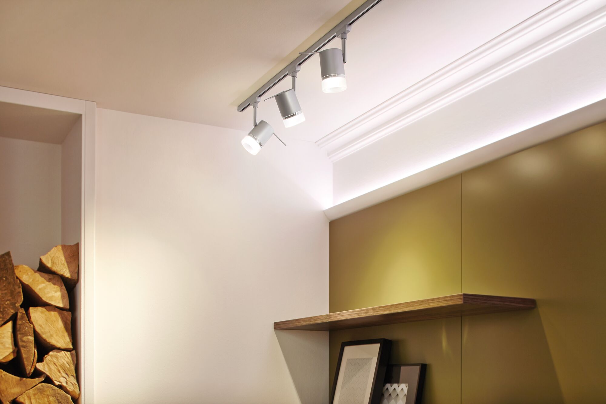 Three white LED spotlights on grey track mounted on ceiling in modern room with wooden shelf and pictures.