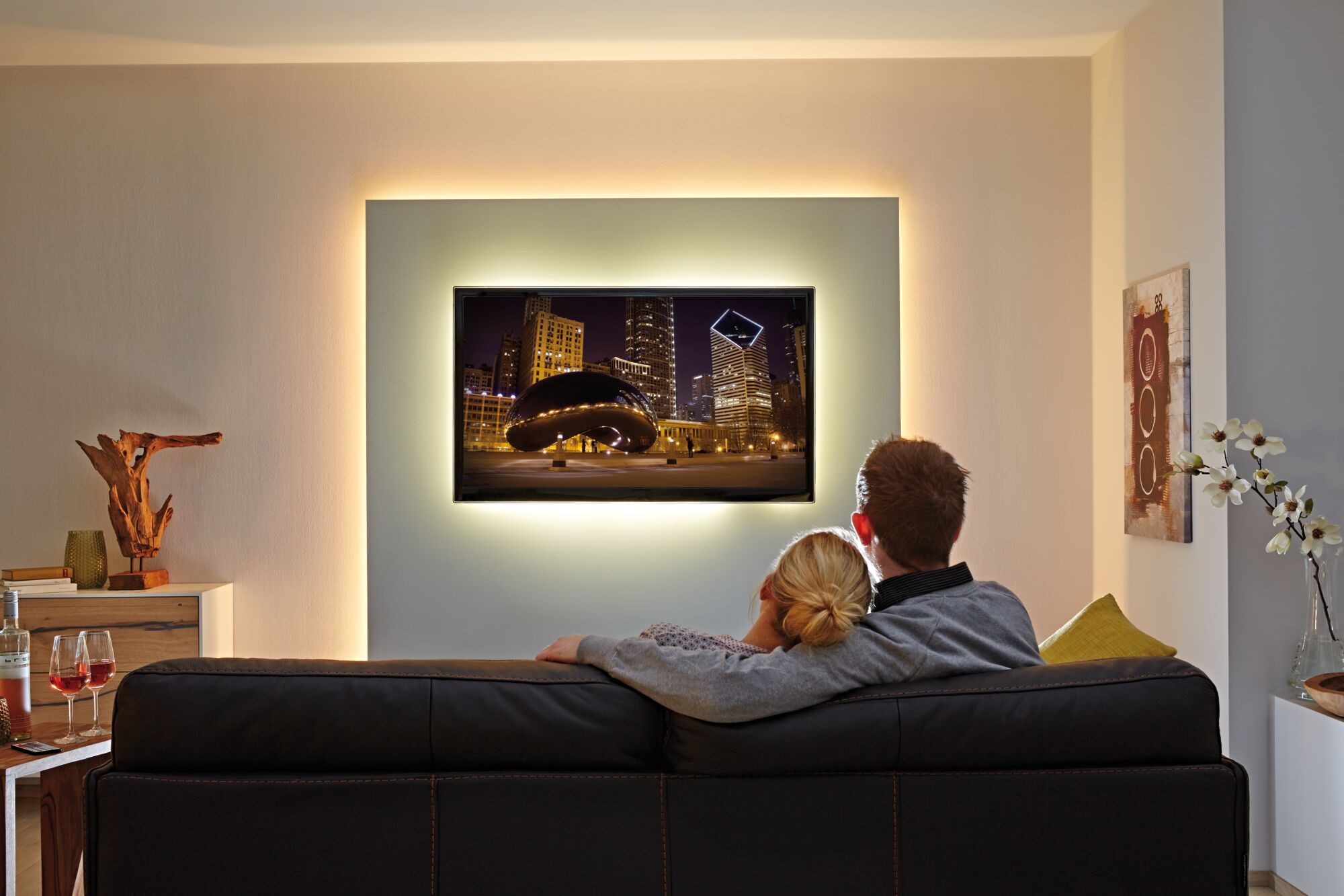 Couple on black leather sofa watching TV with ambient wall lighting in cozy living room