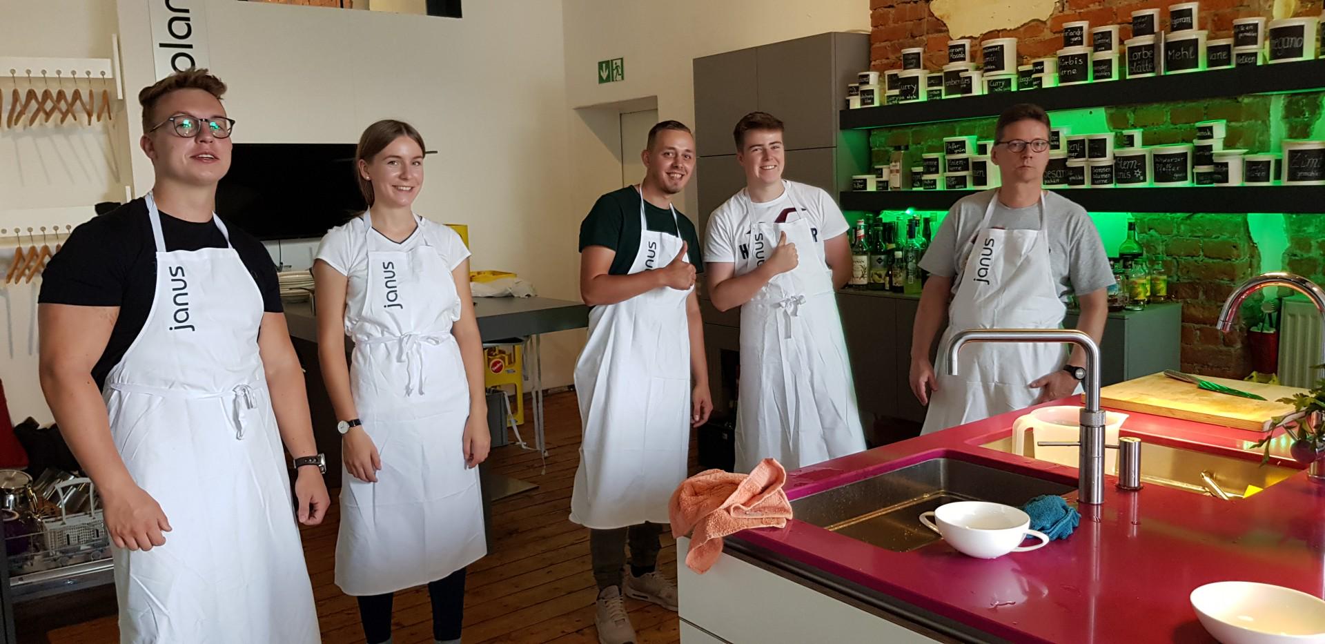 Fünf junge Menschen in weißen Janus-Schürzen in moderner Küche mit roter Arbeitsfläche und Gewürzregal.
