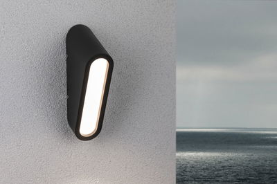 Schwarze LED-Wandleuchte aus Metall mit länglichem Leuchtfeld an weißer Außenwand mit Meerblick