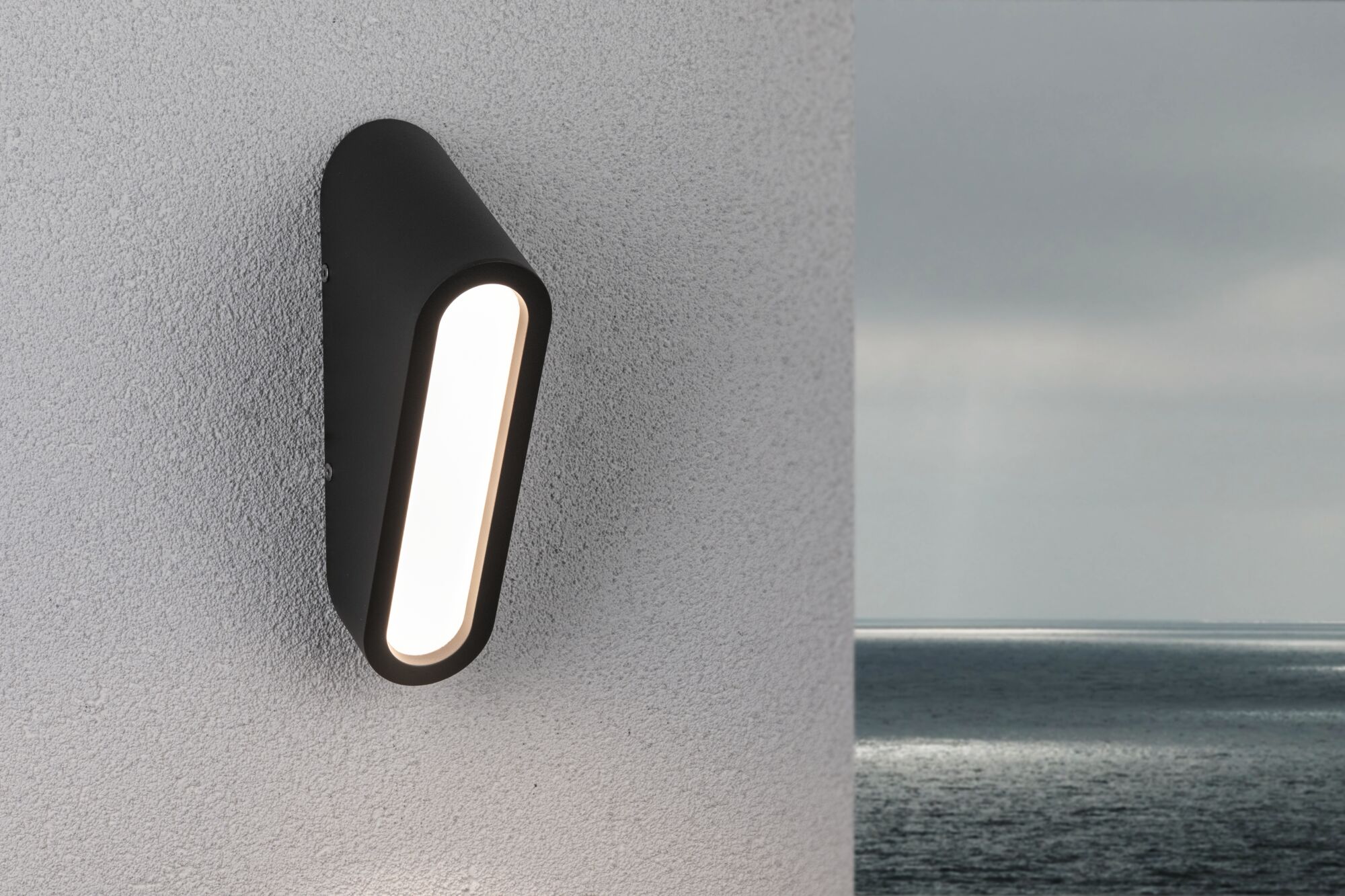 Black metal LED wall light with elongated illuminated panel on white exterior wall by the sea
