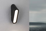 Black metal LED wall light with elongated illuminated panel on white exterior wall by the sea