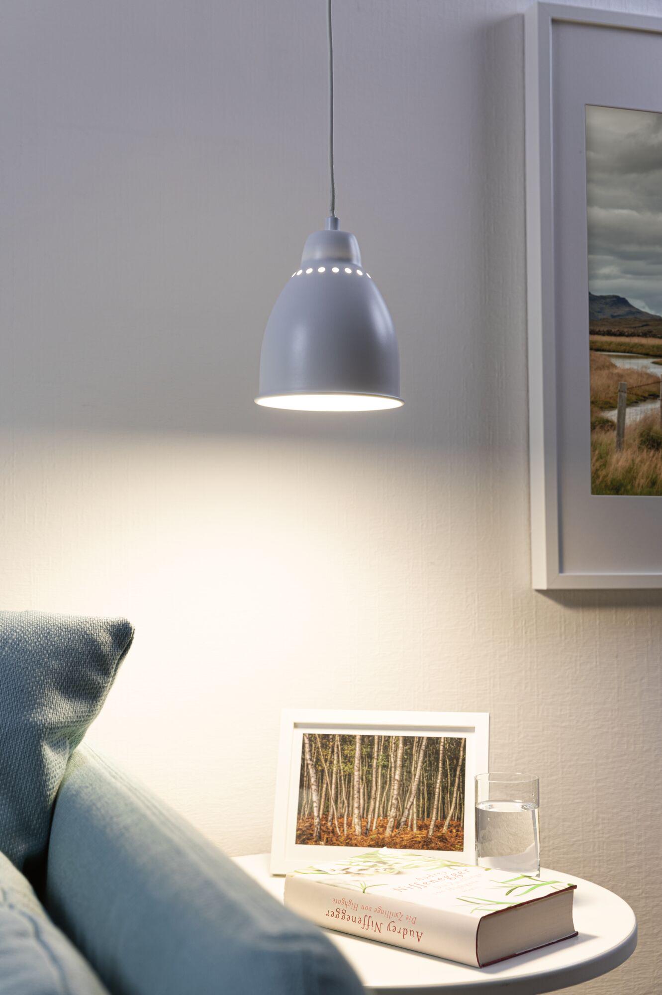 Grey metal pendant lamp with perforated pattern casting warm light over white side table with book and glass.